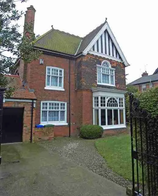 A traditional red brick home with white framed windows and a pointed gable, featuring a gravel driveway and a front yard, behind black metal gates. online homeowner loans.