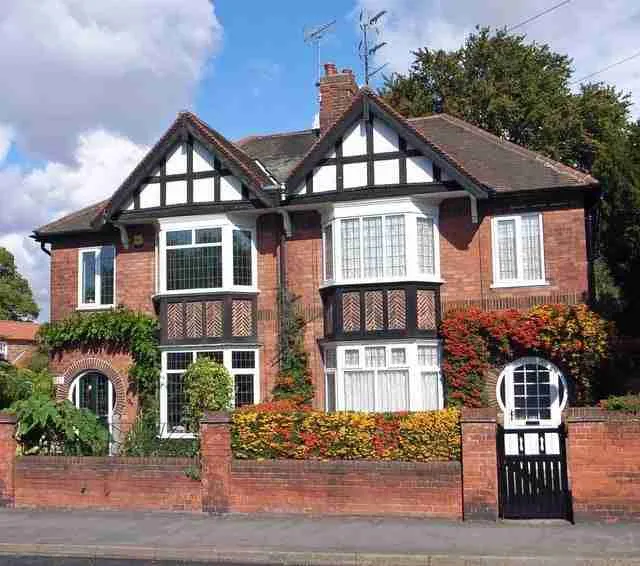A charming two-story brick house with tudor-style half-timbering, featuring a well-manicured garden with colorful flowers and a gated entrance. quick homeowner loans