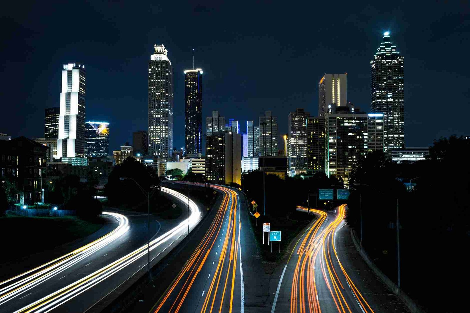 City nightscape with light trails from moving cars on a curving freeway, showcasing a skyline of illuminated high-rise buildings against the dark evening sky.