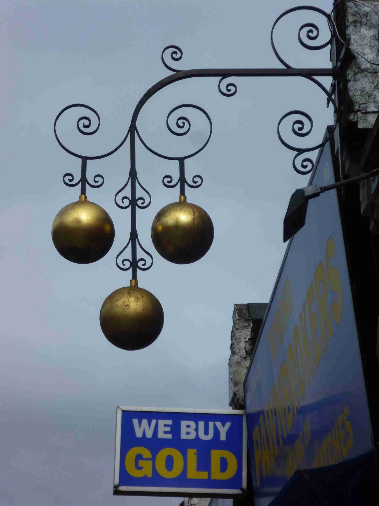 An ornate metal sign with three golden balls hanging against the sky, above a blue 'we buy gold' storefront sign.