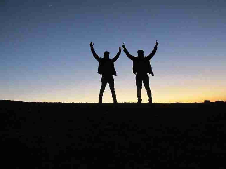 Two silhouetted figures with raised arms against a dusky sky, standing on a horizon line.