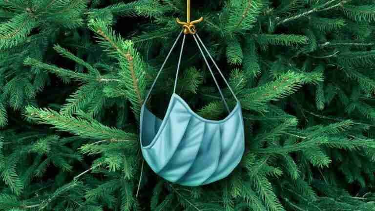 A covid face mask with ear loops hanging from a golden hook, resembling a Christmas ornament, set against the backdrop of dense green fir branches.