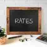 Best Rate Loans With Longer Repayment Periods A wooden-framed chalkboard with the word "RATES" written on it, placed on a desk next to a succulent plant in a decorative pot, a pen, a metallic binder clip, and a calculator. The background is a white brick wall. best rate loans