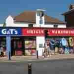 Alt text: A storefront with a sign reading "G&T's Origin Warehouse" selling beach accessories, with people walking in front and a clear blue sky in the background.