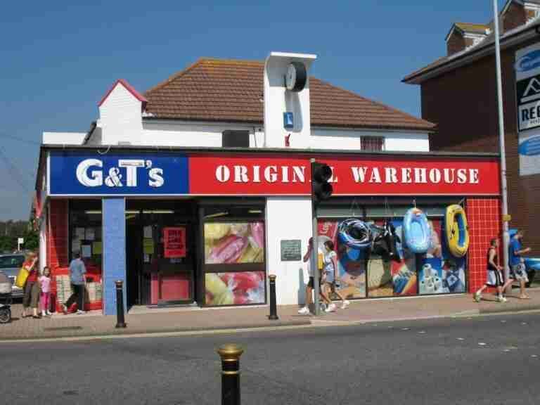 Alt text: A storefront with a sign reading "G&T's Origin Warehouse" selling beach accessories, with people walking in front and a clear blue sky in the background.