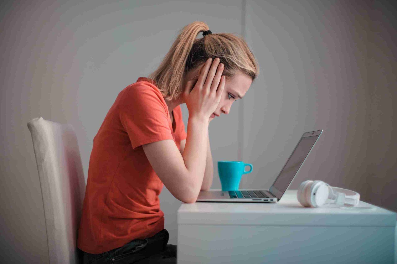 Direct Lender For Bad Credit Loan A woman in an orange t-shirt looks stressed while using a laptop at a white desk with a blue mug and white headphones beside it. direct lender for bad credit loan