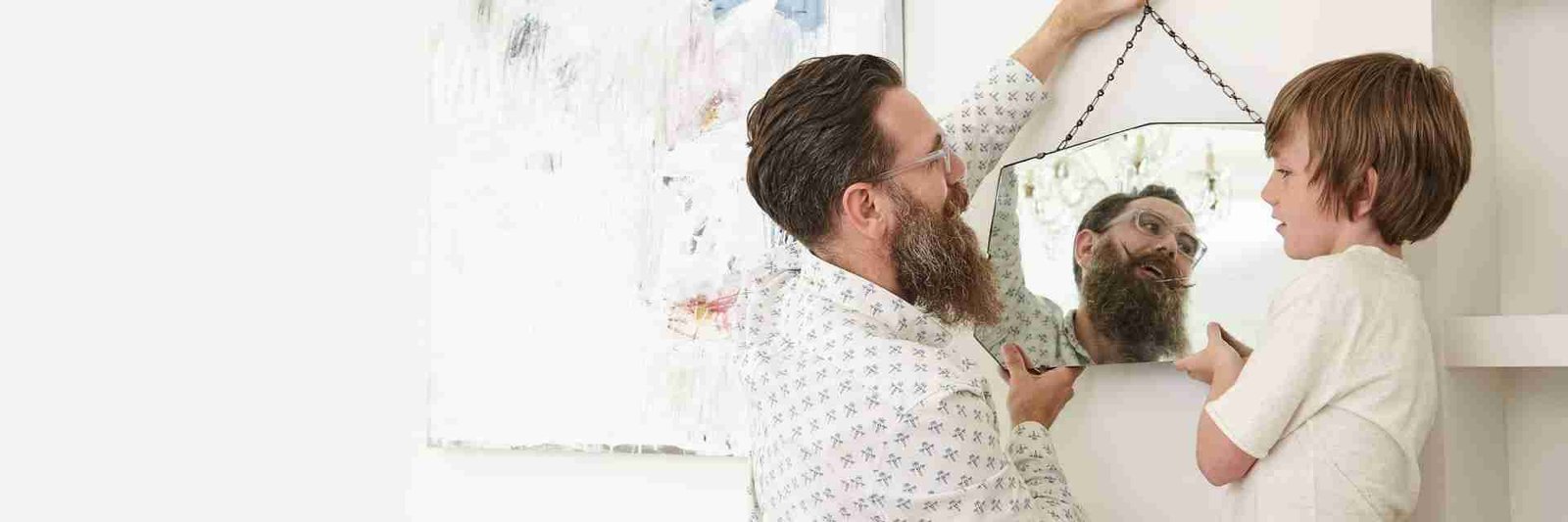 A man with a beard hanging a hexagonal mirror on a wall, assisted by a young boy. They are both looking at the mirror with visible reflection of the man's face. secured loans uk