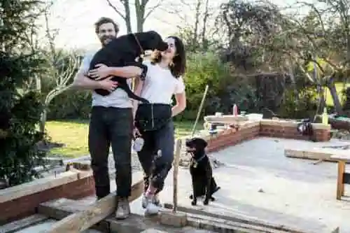 A smiling man holding a large black dog stands beside a woman kissing the dog's face, with another black dog sitting beside them, all on a wooden structure in a garden under construction. best home improvement loans