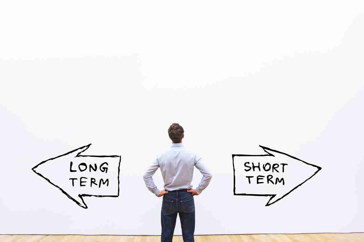 A person standing with hands on hips looking at a wall with two arrows pointing in opposite directions; one arrow labeled "LONG TERM" and the other "SHORT TERM".