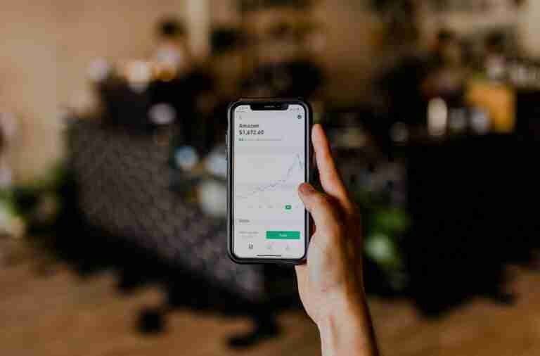 A hand holding a smartphone with a stock trading app open, displaying a rising stock price chart for Amazon. The background is blurred with indistinct features of a room. £100 loans