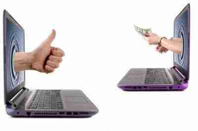 A person giving a thumbs up through one laptop screen to another person offering cash through a second laptop screen, symbolizing an agreement to borrow money now or deal approval.