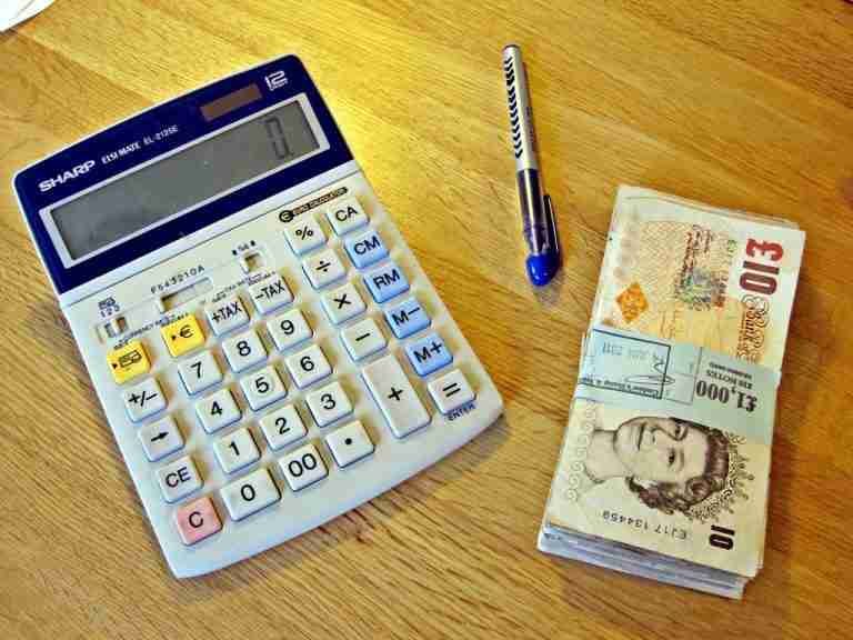 A calculator, a pen, and a stack of British pound banknotes on a wooden surface suggest financial calculations or budgeting for cash advance loans online direct lenders.