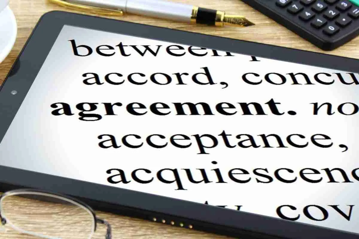 A tablet displaying synonyms for the word "agreement" such as "accord," "concur," and “guaranteed acceptance,” with a pen, glasses, and calculator nearby on a wooden
