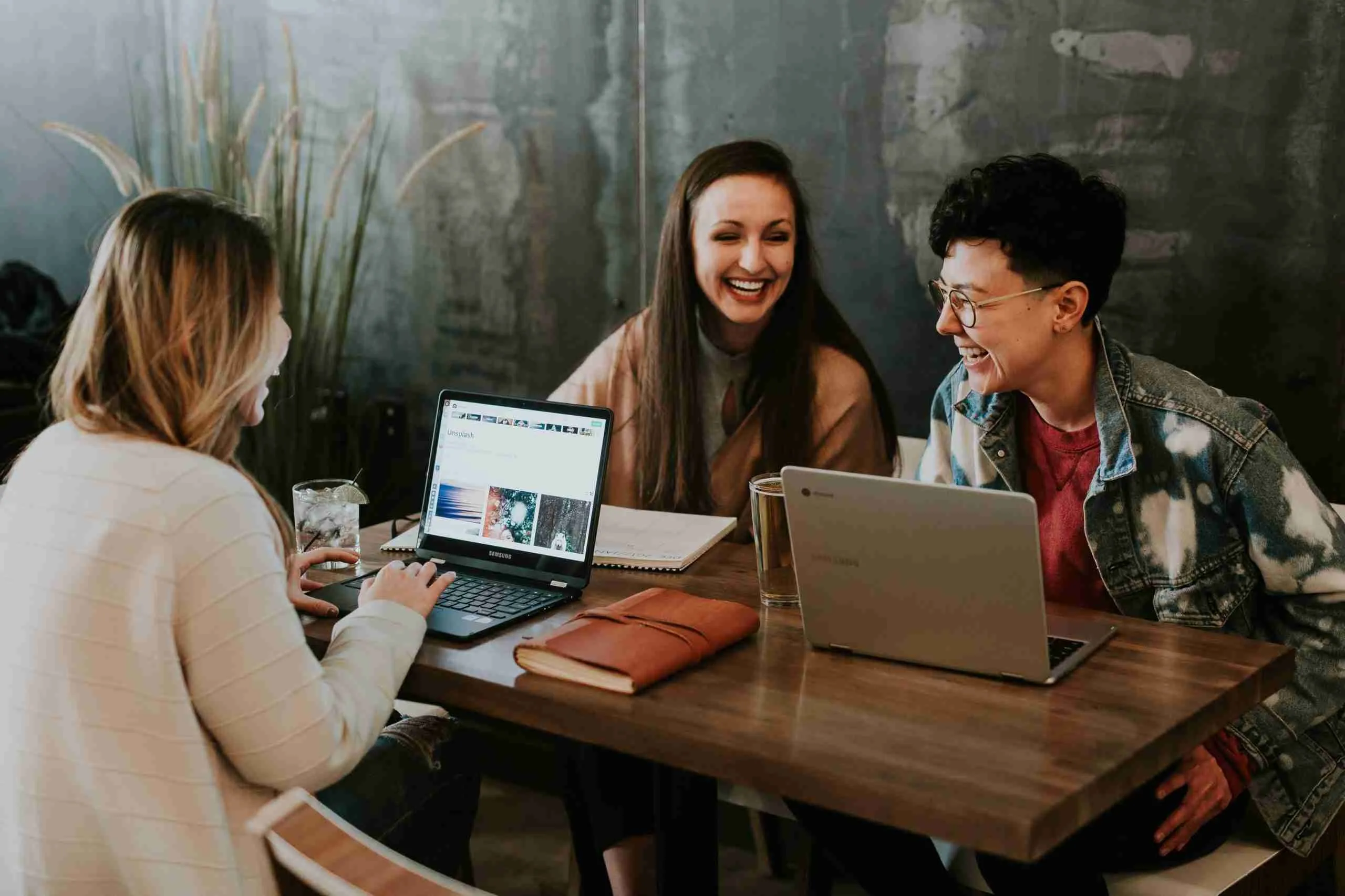 Three friends, discussing loans for students, enjoy a light-hearted moment at a cozy cafe, with laptops suggesting a casual meeting or work session.