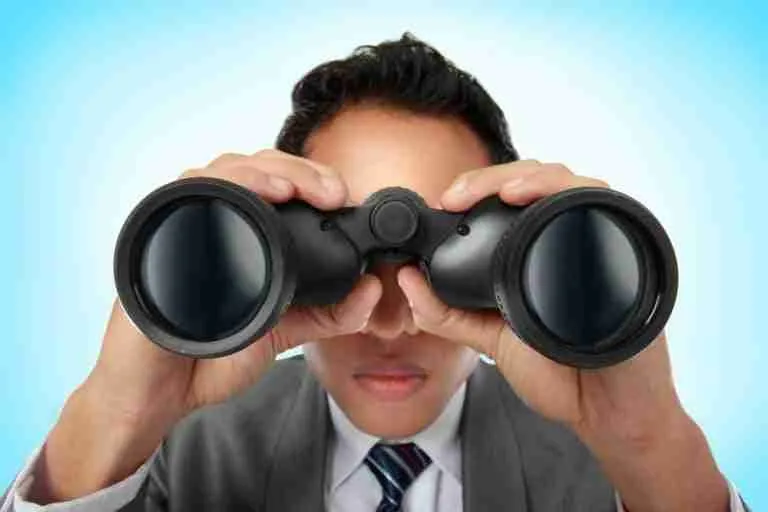 A man in a business suit looking through binoculars with a focused expression, searching for a loan against a light blue background.