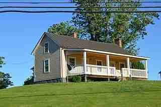 A quaint house with a wraparound porch perched on a green hillside under a clear blue sky offers tenant guarantor loans.